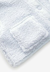 Lichtblauwe fleece kleding met een gestructureerde, fluffy buitenkant, zachte blauwe voering en een klein zakje op de voorkant.