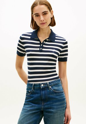 Jeune femme aux cheveux courts bruns portant un polo à manches courtes rayé bleu marine et blanc et un jean en denim bleu sur un fond uni.