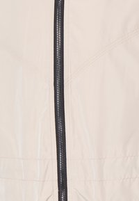 Ljusbeige jacka med en svart dragkedja, slät yta och diskreta sömdetaljer. Materialet verkar slätt och lätt.