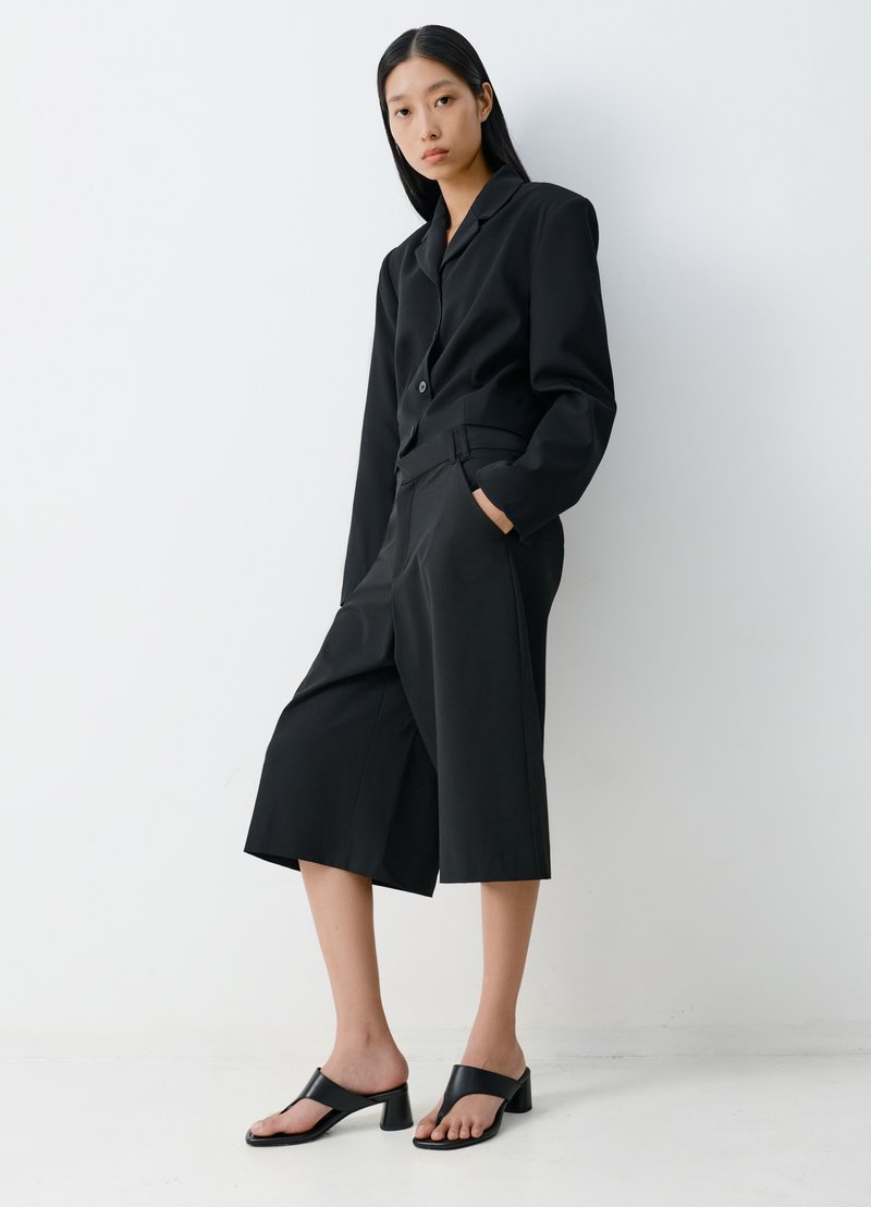 Frau mit langen schwarzen Haaren trägt einen schwarzen Blazer mit langen Ärmeln, schwarze weit geschnittene, knöchellange Hosen und schwarze Sandalen mit Absatz, steht vor einer weißen Wand.