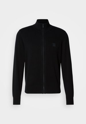 Melnā jaku zupa ar mīkstu adīto audumu, ar stāvošu apkakli, garām piedurknēm un smalku teksturētu logo uzlapu uz krūšu.