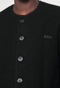 Schwarzer Wollcardigan mit Rundhalsausschnitt, Vorderknöpfen und subtilen Logodetails. Strukturierter Stoff und glatte Knopfakzente.