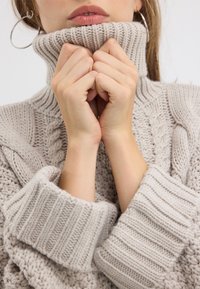 Beige gestrickter Pullover mit dickem geripptem Rollkragen und weiten Bündchen. Verfügt über strukturierte Zopfmuster auf dem Körper.