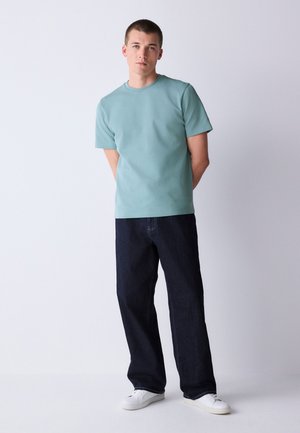 Ung mand står foran en ensfarvet baggrund iført lyseblå kortærmet skjorte, mørke løse jeans og hvide sneakers, hænderne bag ryggen.