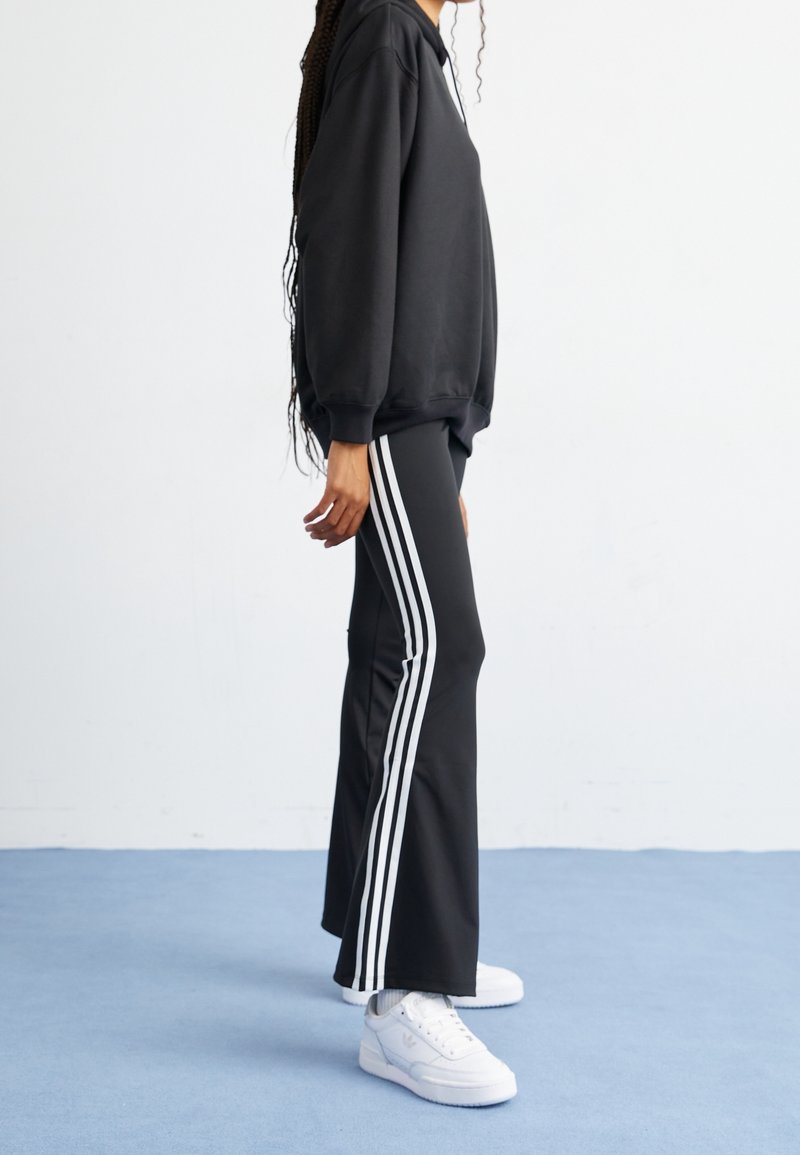 Zwarte sweatshirt met een relaxte pasvorm, gecombineerd met zwarte broek waarop drie witte verticale strepen aan de zijkanten zitten. Witte sneakers maken de look compleet.