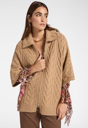 Cardigan beige a lavorazione cable-knit con zip frontale, dotato di colletto e orlo a costine. Indossato sopra una camicetta a fantasia con dettagli floreali.