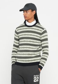 Muškarac s dugom kosom nosi džemper s uzorkom preko bijele košulje, tamnu kapu i tamne trenirke s logotipom "Hollister 22".