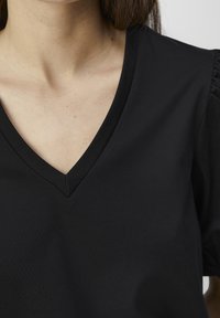 Haut noir à col en V avec manches courtes légèrement bouffantes. Fabriqué en tissu lisse, avec des lignes épurées et un design simple.