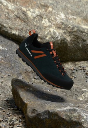 Dunkler Wanderschuh mit orangen Akzenten, der über felsigem Gelände schwebt und den Markennamen Scarpa an der Seite zeigt.