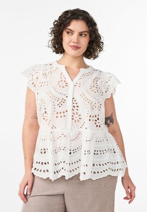 Žena s krátkymi kučeravými vlasmi v bielej čipkovanej blúzke s kvetinovými výrezmi a béžových nohaviciach stojaca pred jednoduchým pozadím.