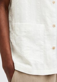 Brazo descansando junto a una camisa blanca texturizada con bolsillo delantero y pantalones beige, mostrando detalles de la tela y acentos de botones.