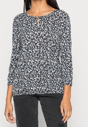 Mujer con camiseta negra de manga larga con un patrón moteado blanco y marrón y un pequeño escote en forma de ojo de cerradura, combinada con pantalones negros.