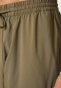 Olīvu zaļie sporta bikses ar elastīgu jostasvietu un auklu. Gluda, viegla auduma struktūra ar nelielu spīdumu un brīvu piegriezumu.