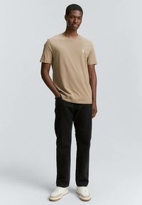 Beżowy bawełniany t-shirt z krótkimi rękawami i małym logo na piersi, w połączeniu z czarnymi jeansami i białymi trampkami. Jednolite tło.