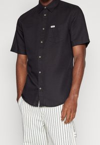 Chemise noire à manches courtes avec des boutons, une poche poitrine et une petite étiquette de marque. La texture du tissu semble lisse, offrant un design décontracté.