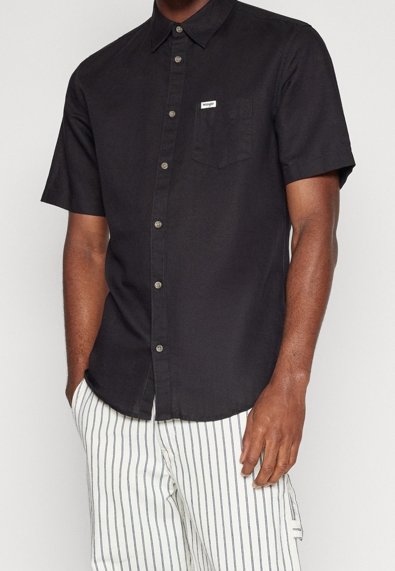 Chemise noire à manches courtes avec des boutons, une poche poitrine et une petite étiquette de marque. La texture du tissu semble lisse, offrant un design décontracté.