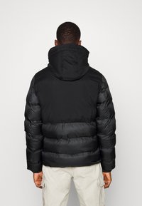 Sort polstret jakke med hætte, der har en glat øverste sektion og en fyldig nedre sektion. Stoffet ser skinnende og isoleret ud.