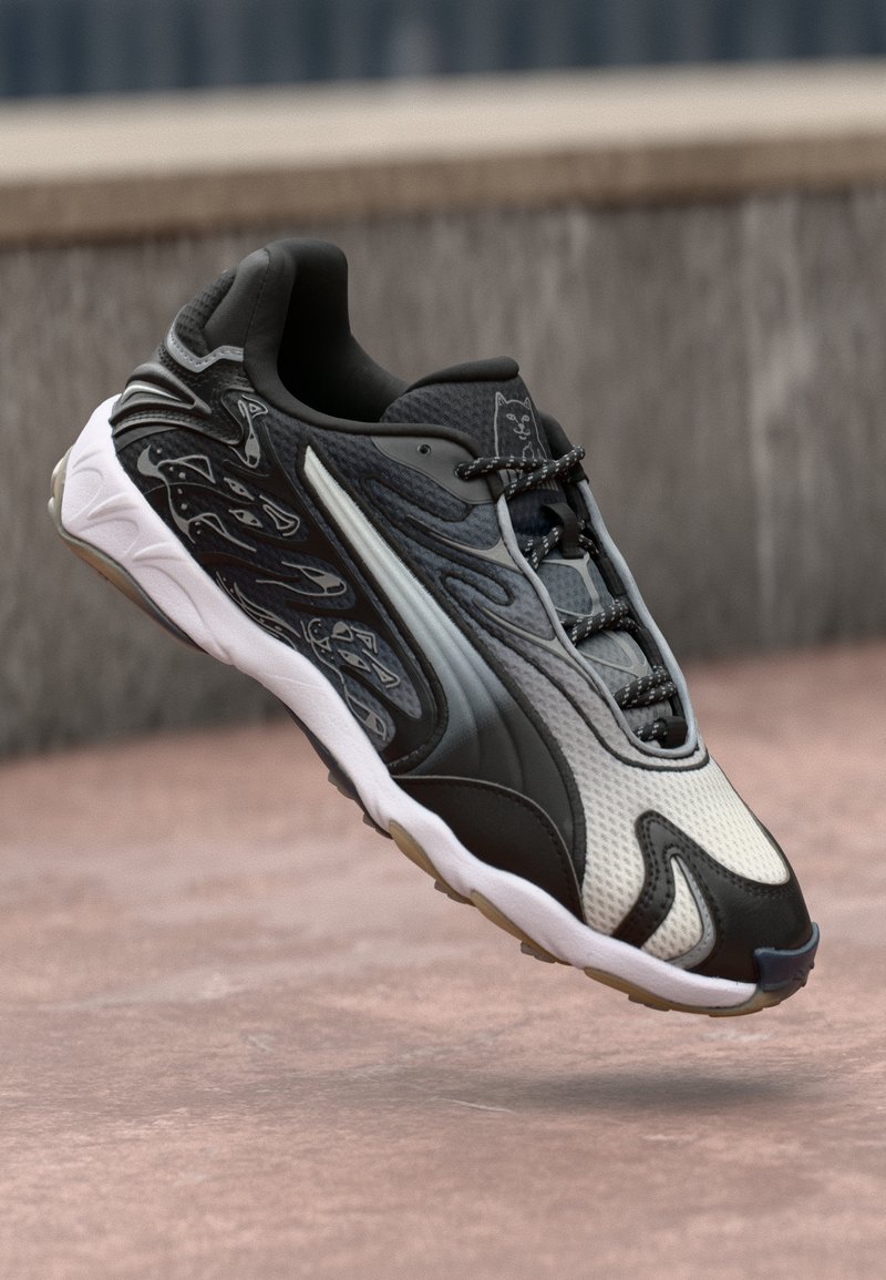 Chaussure de sport noire et blanche avec des motifs abstraits, visage de chat sur la languette et semelle blanche incurvée, sur un fond urbain flou.