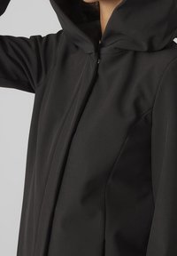 Chaqueta negra con capucha hecha de tela suave, con cremallera frontal y mangas estructuradas y entalladas, con un diseño minimalista.