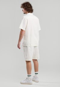 Camicia beige a maniche corte e pantaloni corti abbinati in tessuto leggero. Il modello indossa sneakers bianche con calze a righe verdi.