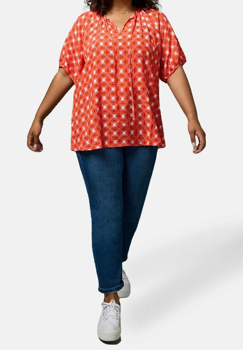 Red geometric-patterned blouse with a V-neckline and short, wide sleeves, paired with dark blue jeans and white sneakers.