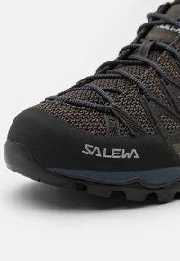 Tamsiai rudos spalvos lauko batai su tinklo viršumi, guminėmis pirštų galūnėmis ir tekstūruotomis raištelėmis. Šone yra "SALEWA" logotipas ir tvirtas pado dizainas.