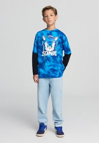 Maglietta tye-dye blu con grafica di Sonic the Hedgehog, abbinata a jeans azzurri e sneakers blu scuro. Maniche lunghe nere visibili.