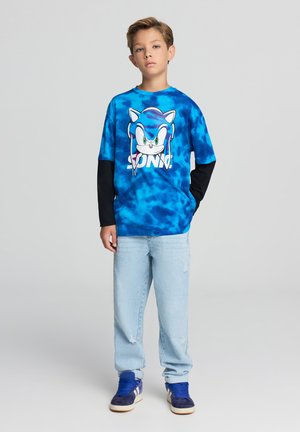 Maglietta tye-dye blu con grafica di Sonic the Hedgehog, abbinata a jeans azzurri e sneakers blu scuro. Maniche lunghe nere visibili.