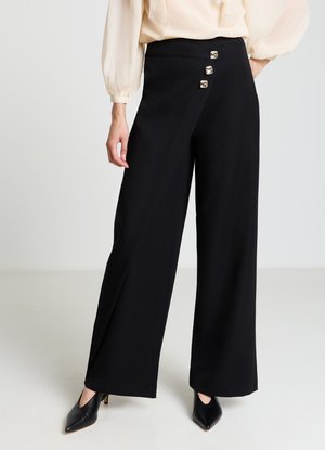 Pantalon noir à jambes larges avec trois boutons métalliques sur la taille gauche. Tissu lisse, assorti à un chemisier léger et transparent ainsi qu'à des chaussures à bout pointu.