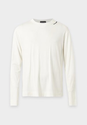 Giovane uomo in piedi che indossa una camicia bianca a maniche lunghe, jeans azzurri a gamba larga e scarpe da ginnastica bianche su uno sfondo semplice.