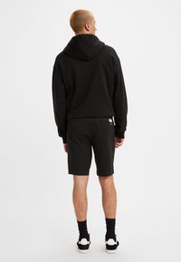 Svart hettegenser med hette, kombinert med svarte shorts. Viser bakvinkel, med teksturert stoff, tettsittende design og avslappede joggesko.