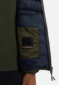 Maniche della giacca a vento in blu navy e oliva, con tasca a toppa dotata di un quadrato nero e logo, e cordoncino regolabile al polsino.