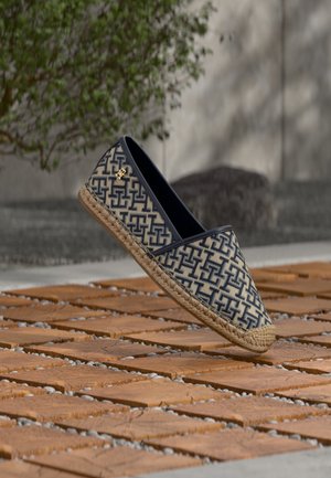 Kék és bézs mintás espadrille fonott juta talppal, egy fa csempepadló felett lebegve, háttérben zöld növényzettel.