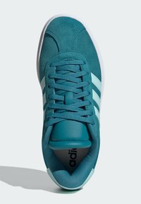 Teal mockasin med vit gummisula, med tre aqua-ränder, rund tå och texturerade snören. Ren, minimalistisk design.