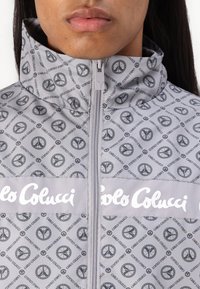Grijze zip-up jack met een patroon van overal verspreide vredessymbolen, met een hoge kraag en het "Carlo Colucci" logo in het wit over de borst.
