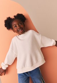 Felpa in tessuto morbido bianco, vestibilità oversize con spalle scese. Abbinata a jeans di denim blu. Sfondo con accento color pesca.