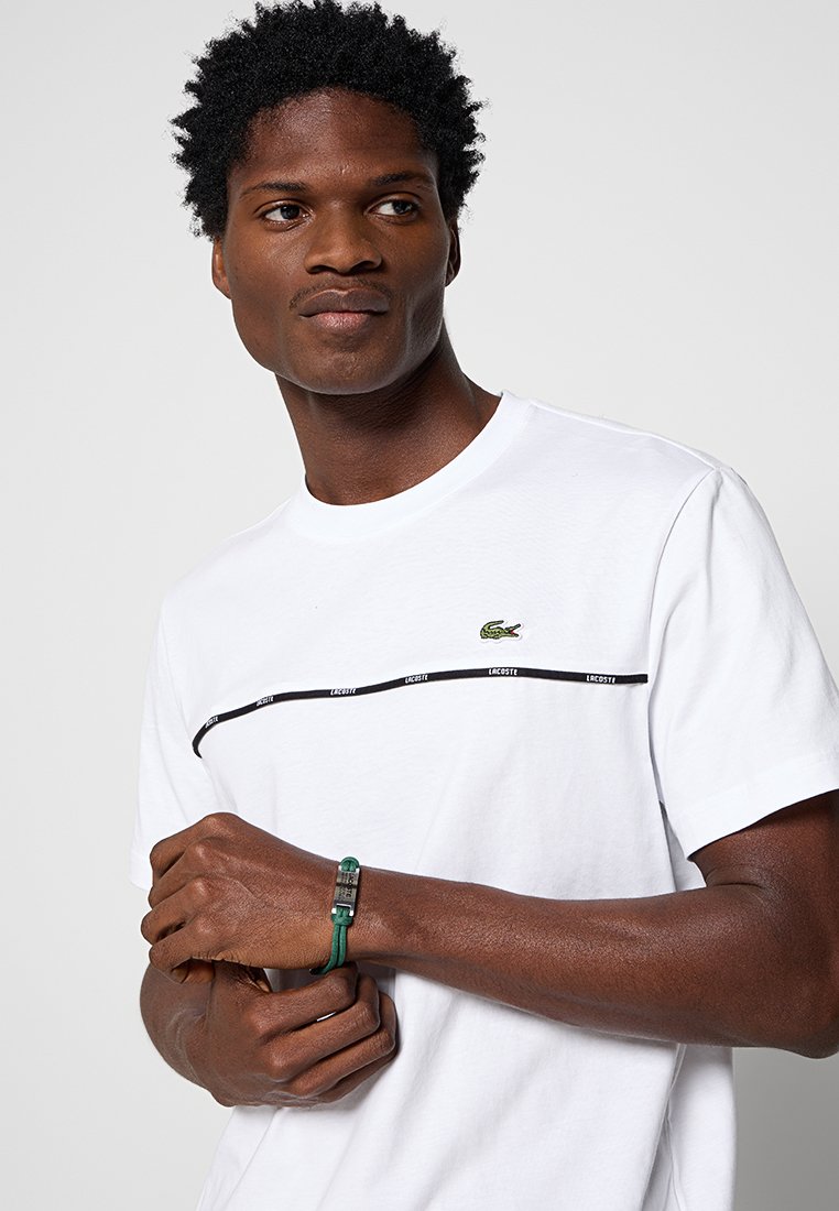 Weißes Baumwoll-T-Shirt mit schwarzem horizontalem Streifen, auf dem "LACOSTE" steht, grünes Krokodil-Logo auf der Brust, getragen mit einem grünen Armband.