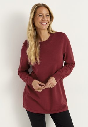 Kvinde med langt blondt hår, der smiler, iført en langærmet vinrød sweater og sorte bukser, stående foran en ensfarvet lys væg.