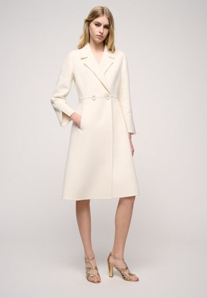 Cream-colored wool coat with a tailored design, double-breasted buttons, lapel collar, and flared sleeves. Paired with strappy gold heels.