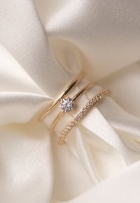 Trois bagues en or sur un satin blanc : l'une avec un diamant central, une autre avec de petites pierres transparentes, et une simple bande fine.