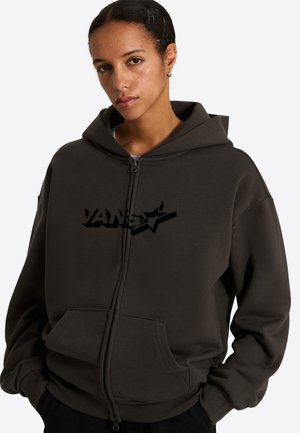 Young woman wearing a dark zip-up hoodie with a hood and a black Vans logo, hands in pockets, looking at the camera.
