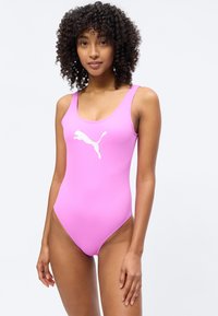 Γυναίκα με σγουρά μαλλιά που φοράει ένα ροζ μαγιό χωρίς μανίκια με λευκό λογότυπο Puma, στέκεται μπροστά σε απλό φωτεινό φόντο.