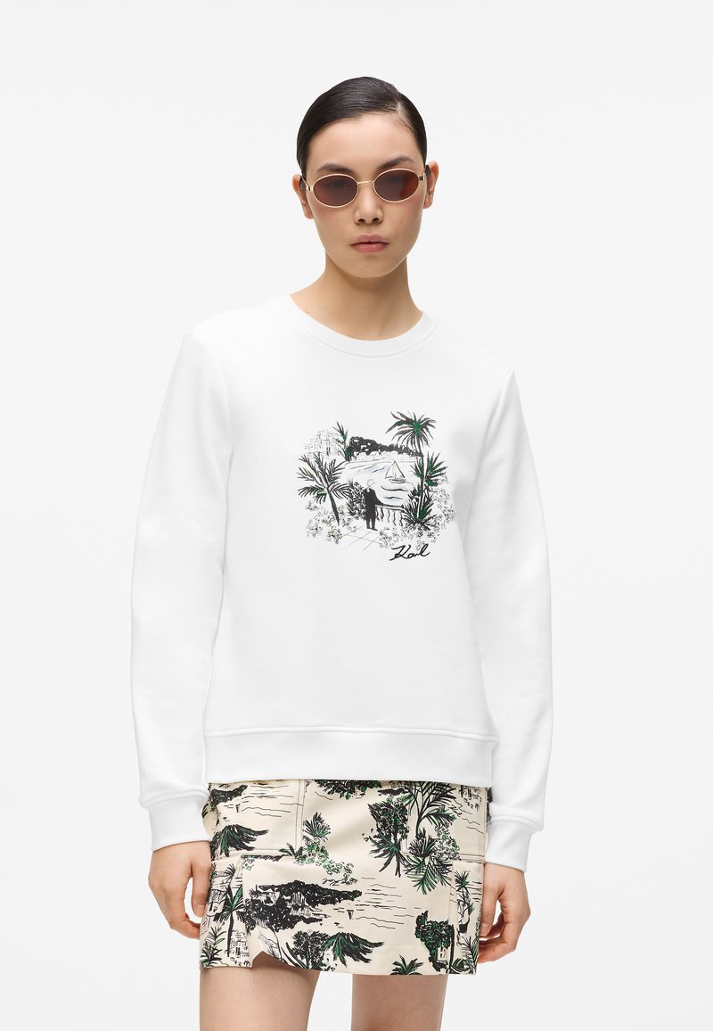 Sweat-shirt blanc avec un imprimé graphique tropical noir et vert, associé à une jupe à motifs avec des motifs de plantes en beige et tons foncés.
