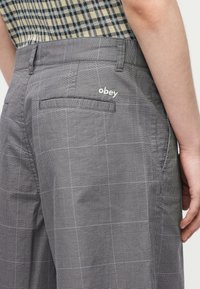 Κοντινό πλάνο ενός ατόμου που φοράει γκρι καρό παντελόνι με κεντημένο λογότυπο "obey" πάνω από την πίσω τσέπη και πουκάμισο καρό μεσαίο μέσα.