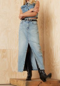 Denim maxikjol, ljusblå, med en framtida slits. Matchad med svarta knähöga stövlar. Outfiten består av en kort denim topp och ett svart bälte.