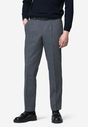 Man wearing grey tailored trousers, black woven belt, black leather loafers, and a dark long-sleeve sweater, standing with hands partially in pockets.