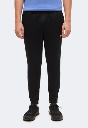 Muž nosí čierne Nike tepláky s malým bielym logom na pravom stehne a čierne športové topánky.