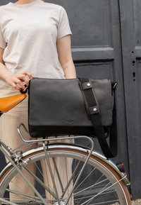 Svart läder budväska med avtagbar axelrem, med silverdetaljer och en slät textur, placerad på en cykel.