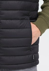 Czarna kamizelka puchowa z pikowanym, wypełnionym wzorem. Posiada boczną kieszeń z zamkiem błyskawicznym oraz metkę oznaczoną "BLENDO". Zielony sweter w prążki pod spodem.