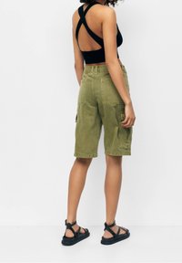 Olijfgroene cargoshorts met een relaxed fit, voorzien van zijzakken en een gladde textuur. Gemaakt in combinatie met een zwart topje met kruisbanden op de rug en zwarte sandalen.